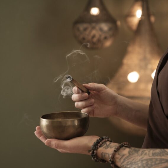 Räucher-Ritual: Eine Person mit einer Klangschale aus Messing, aus der Weihrauch aufsteigt, mit Perlenarmbändern und Tattoos, mit warmen Lichtern im Hintergrund.
