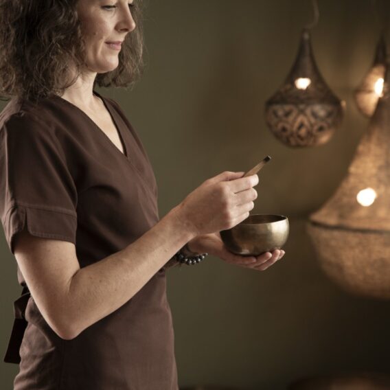 Räucher-Ritual: Eine Frau in einer braunen Uniform hält eine Klangschale in einem warm beleuchteten Raum mit dekorativen Hängelampen.