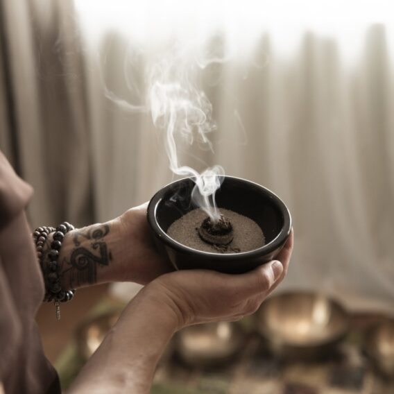 Eine Person, die eine Räucherschale mit Sand in der Hand hält, Armbänder und eine Tätowierung trägt, in einem schwach beleuchteten Raum.