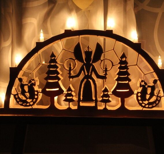 Adventsmarkt im Flürchen: Hölzerner Schwibbogen mit einem Engel, Bäumen und Figuren, beleuchtet von elektrischen Lichtern, vor einem gemusterten Vorhang.
