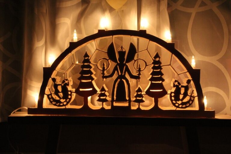 Adventsmarkt im Flürchen: Hölzerner Schwibbogen mit einem Engel, Bäumen und Figuren, beleuchtet von elektrischen Lichtern, vor einem gemusterten Vorhang.