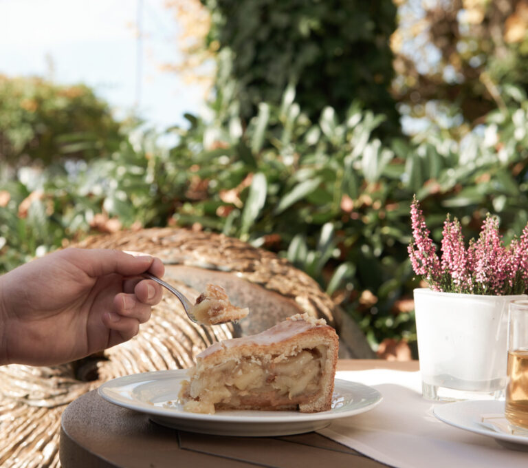 Kaffee & Kuchen: Eine Person hält eine Gabel mit einem Bissen Apfelkuchen an einem Tisch im Freien, mit Pflanzen und einer Topfblume im Hintergrund.