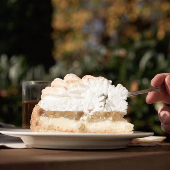 Kaffee & Kuchen: Eine Hand hält eine Gabel, um ein Stück Sahnetorte auf einem Teller im Freien zu essen, mit unscharfem Laub im Hintergrund.