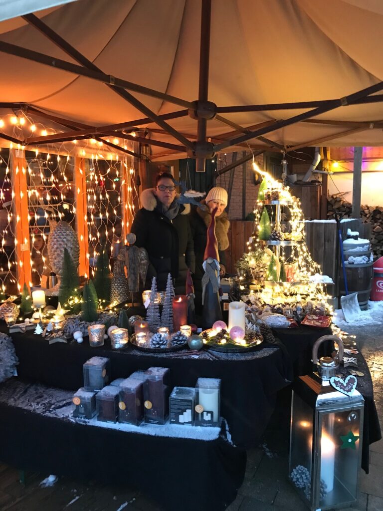 Adventsmarkt im Flürchen: Ein festlicher Marktstand im Freien mit Kerzen, Dekorationen und Lichtern; zwei Personen stehen hinter der Auslage unter einem Vordach.