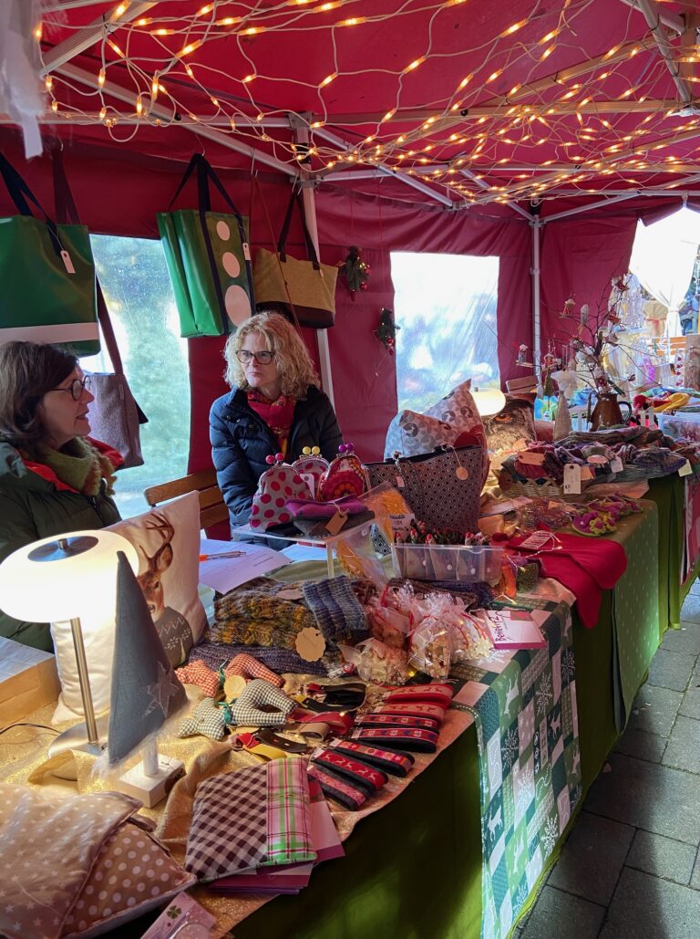 Adventsmarkt im Flürchen: Zwei Frauen sitzen an einem festlichen, mit Lichtern geschmückten Marktstand und verkaufen auf einer grünen Tischdecke handgefertigtes Kunsthandwerk, Taschen und Geschenke.