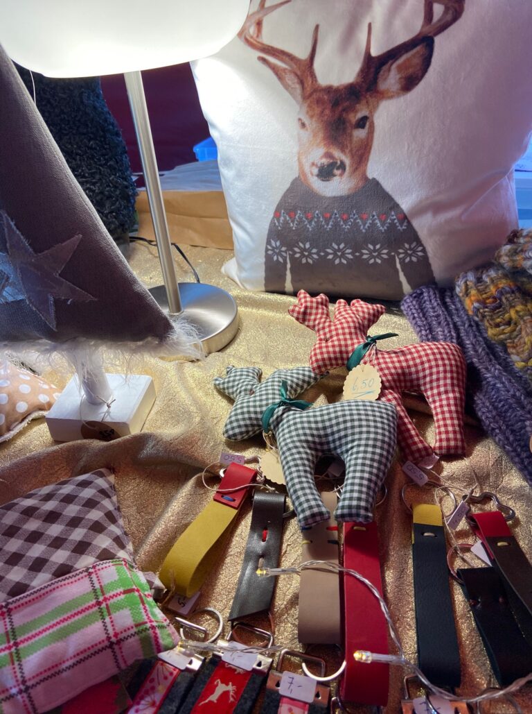 Adventsmarkt im Flürchen: Handgefertigtes Kunsthandwerk wird ausgestellt, darunter Rentiere aus kariertem Stoff, Schlüsselanhänger und ein Kissen mit einem Hirschaufdruck in einem Pullover.