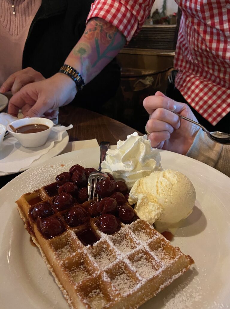 Adventsmarkt im Flürchen: Ein Teller mit einer Waffel, die mit Kirschen, Eis und Schlagsahne belegt ist; am Tisch sitzen zwei Personen, eine davon in einem rot karierten Hemd.
