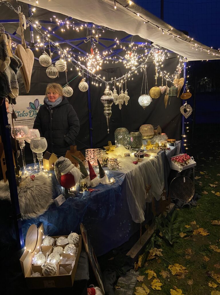 Adventsmarkt im Flürchen: Eine Frau steht an einem festlichen, mit Lichtern geschmückten Marktstand und verkauft an einem dunklen Abend Schmuck und Kunsthandwerk für die Feiertage.