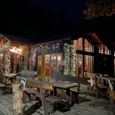 Adventsmarkt im Flürchen: Holztische vor einem gemütlichen, nachts mit Lichterketten geschmückten Restaurant; warmes Licht leuchtet von innen.