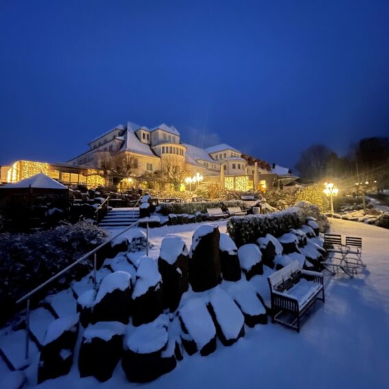 Ein großes Gebäude mit warmen Lichtern erstrahlt inmitten des verschneiten Geländes und der Sitzplätze im Freien in einer Winternacht.