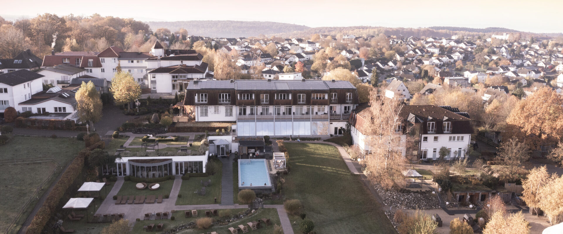 Luftaufnahme eines Hotels mit Gärten, einem Pool und Liegestühlen, umgeben von Häusern und Herbstbäumen bei Sonnenuntergang.