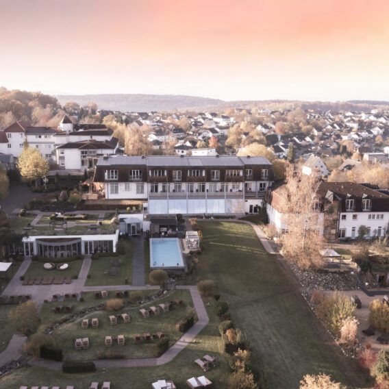 Luftaufnahme eines Hotels mit Gärten, einem Pool und Liegestühlen, umgeben von Häusern und Herbstbäumen bei Sonnenuntergang.