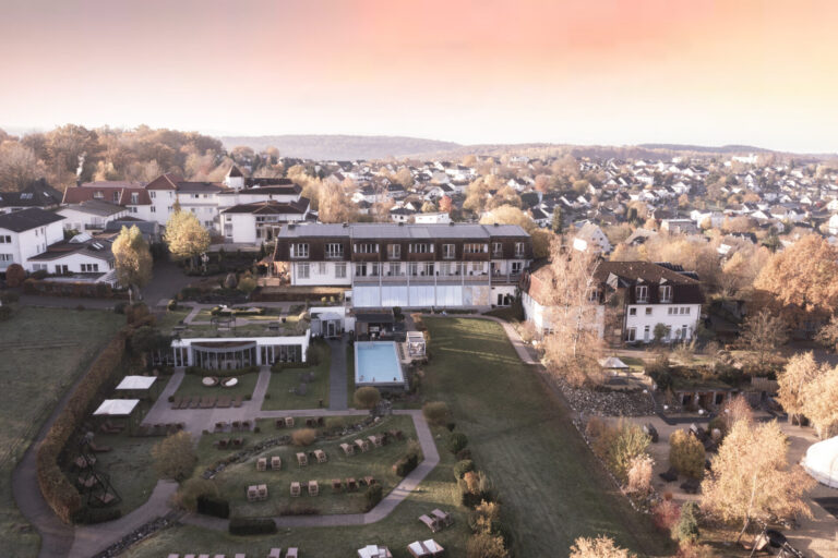 Luftaufnahme eines Hotels mit Gärten, einem Pool und Liegestühlen, umgeben von Häusern und Herbstbäumen bei Sonnenuntergang.