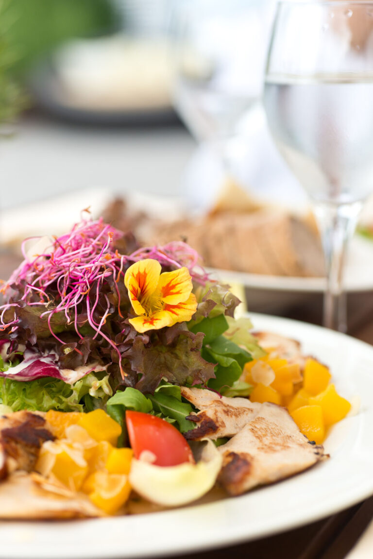 Nahaufnahme eines frischen Salats mit gegrilltem Hähnchen, buntem Grünzeug, essbaren Blüten, gewürfelten gelben Paprikaschoten und einem Glas Wasser im Hintergrund.