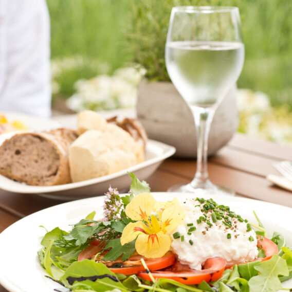 Salatteller mit Gemüse, Tomaten, essbaren Blüten und Käse, daneben Brot und ein Glas Wasser auf einem Tisch im Freien.