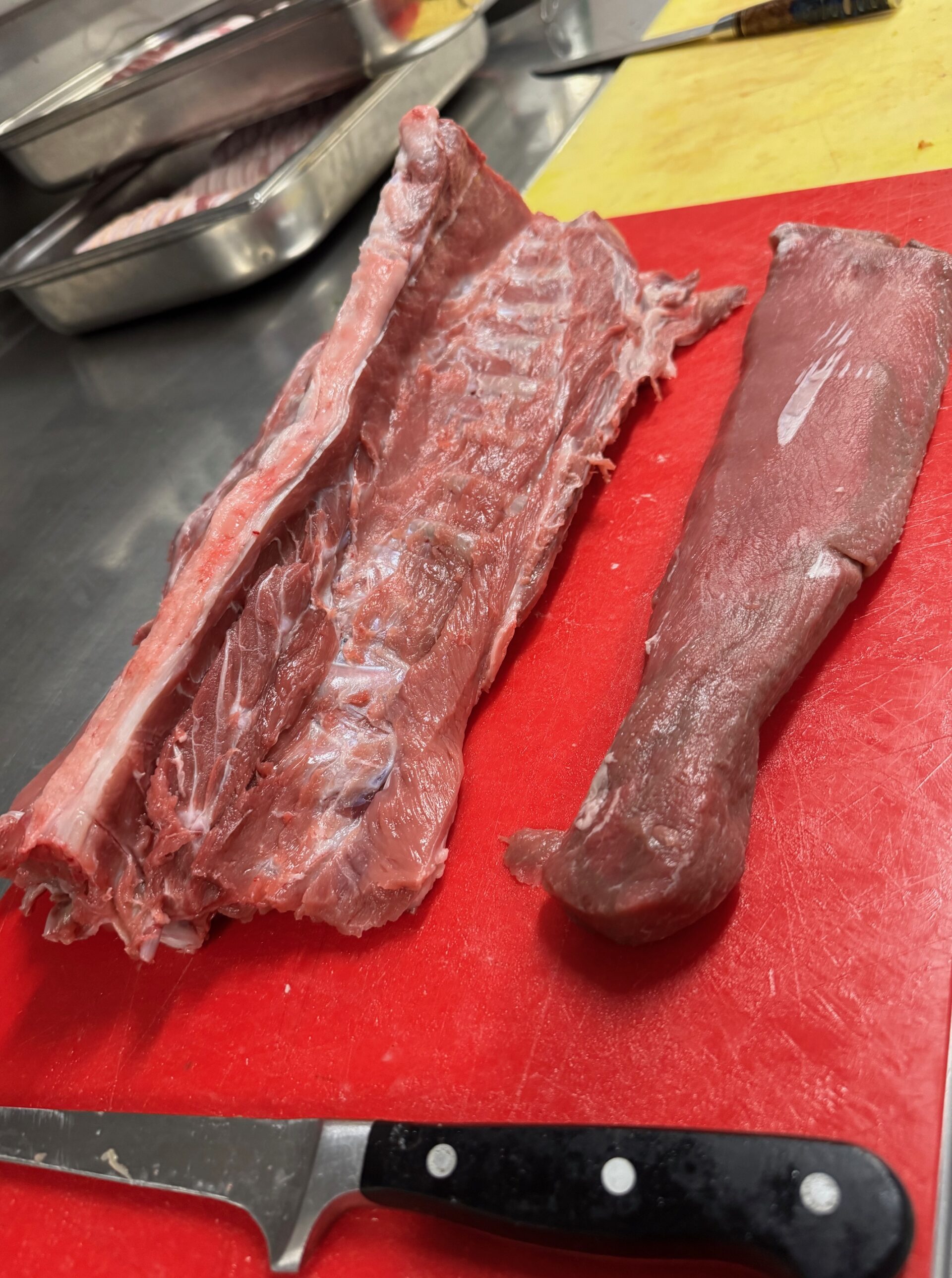 Rohe Fleischstücke und ein Messer auf einem roten Schneidebrett, mit Metallschalen und einem gelben Schneidebrett im Hintergrund.