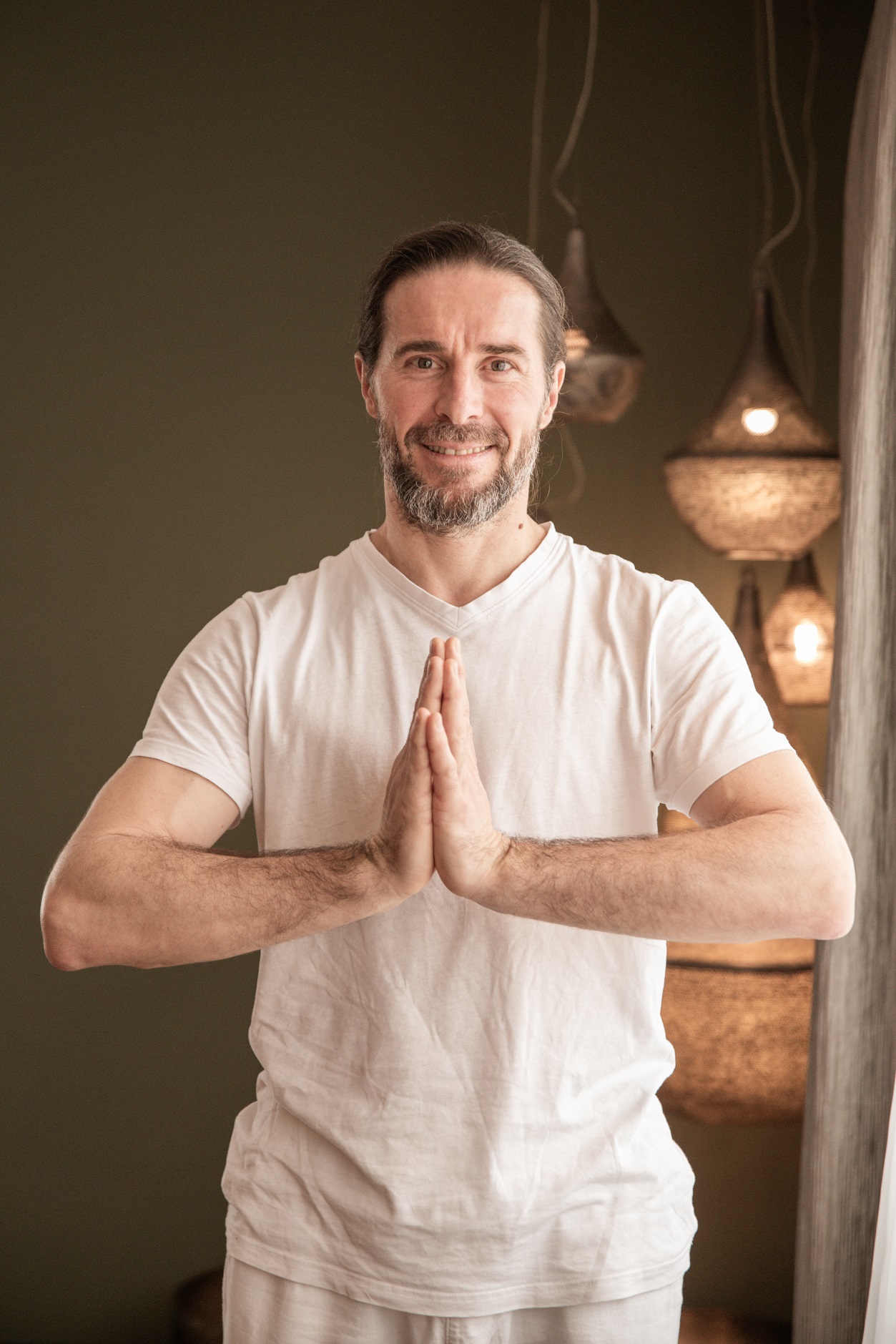 Ein Mann in einem weißen Hemd steht drinnen und lächelt, die Hände in einer Yoga- oder Gebetshaltung zusammengepresst.