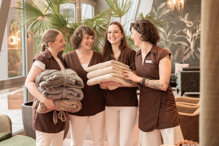 Vier Frauen in braunen Uniformen, lächelnd und mit Handtüchern und Decken in der Hand, in einer gemütlichen, an ein Spa erinnernden Umgebung mit Pflanzen im Hintergrund.