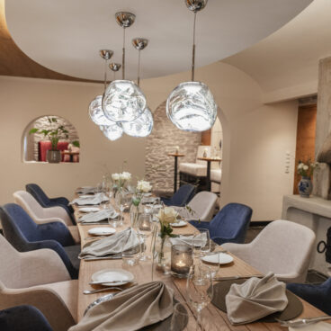 Ein modernes Esszimmer mit einem langen Holztisch, der für eine Mahlzeit gedeckt ist, eleganten blauen und beigen Stühlen und einzigartigen Pendelleuchten über dem Kopf.