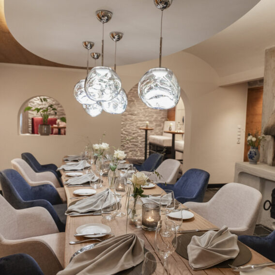 Ein modernes Esszimmer mit einem langen Holztisch, der für eine Mahlzeit gedeckt ist, eleganten blauen und beigen Stühlen und einzigartigen Pendelleuchten über dem Kopf.