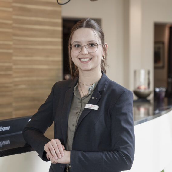 Eine Frau im Anzug und mit Namensschild steht lächelnd an der Rezeption in einer modernen, gut beleuchteten Lobby.